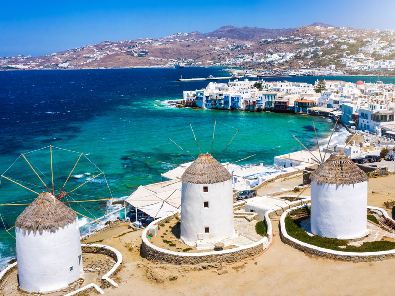 Tre hvite vindmøller har utsikt over en turkis bukt og en hvitkalket landsby på en solrik dag. Mykonos, Hellas