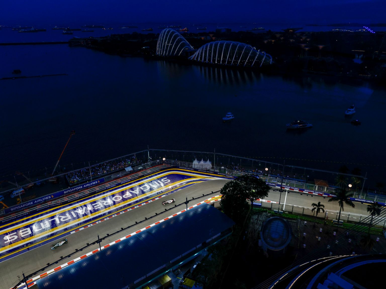 Éjszakai légi felvétel a Szingapúri F1 pályáról versenyautókkal, kivilágított Gardens by the Bay-jel és hajókkal az öbölben.
