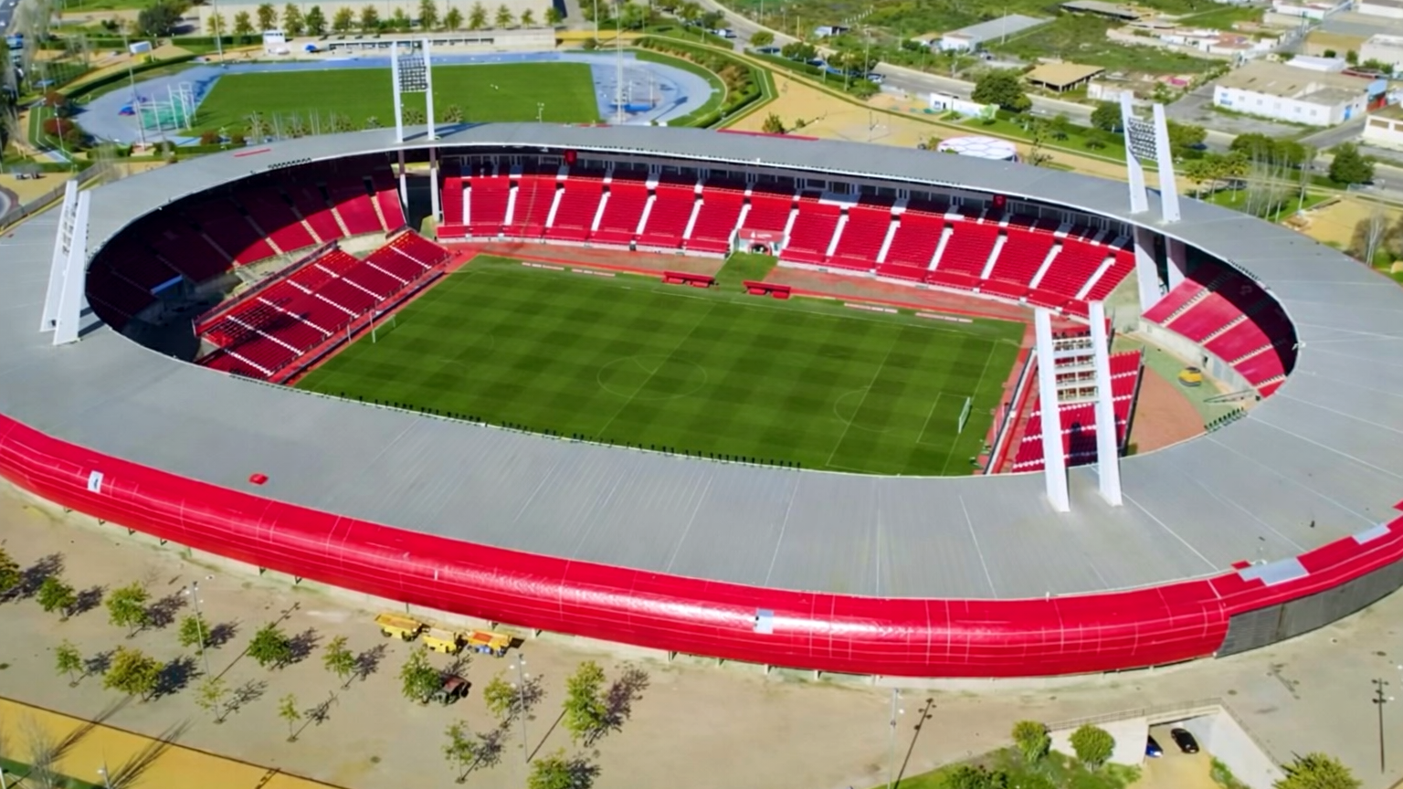 Vista aérea de un gran estadio con asientos rojos y un campo verde, con un techo gris y paredes exteriores rojas. Un estadio más pequeño es visible al fondo.