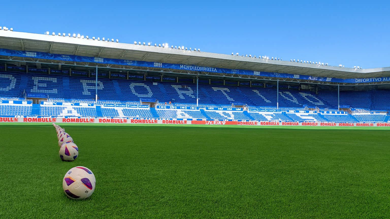Futballstadion zöld pályával, kék ülésekkel és egy sor futball-labdával tiszta ég alatt.