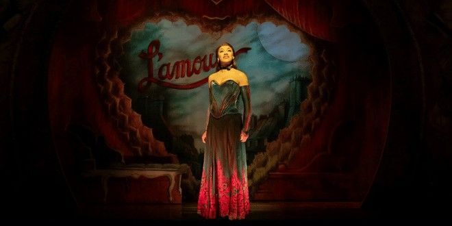 Eine Frau in einem dunkel petrolfarbenen und roten Kleid steht auf einer Bühne vor einem Hintergrund, auf dem das Wort „L'amour“ in einem Herz zu sehen ist.