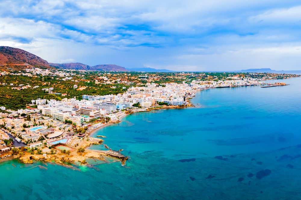 Luftfoto af en kystby med hvide bygninger, strande og bjerge under en overskyet himmel.