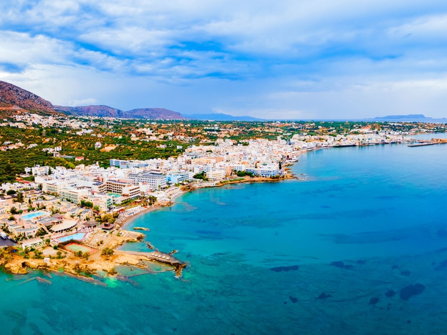 Luftfoto af en kystby med hvide bygninger, strande og bjerge under en overskyet himmel.