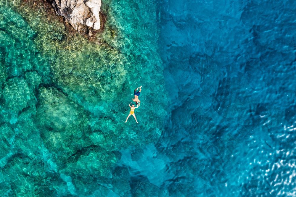 Aerial view of two people floating in clear blue-green ocean water near a rocky coastline. Rhodes, Greece