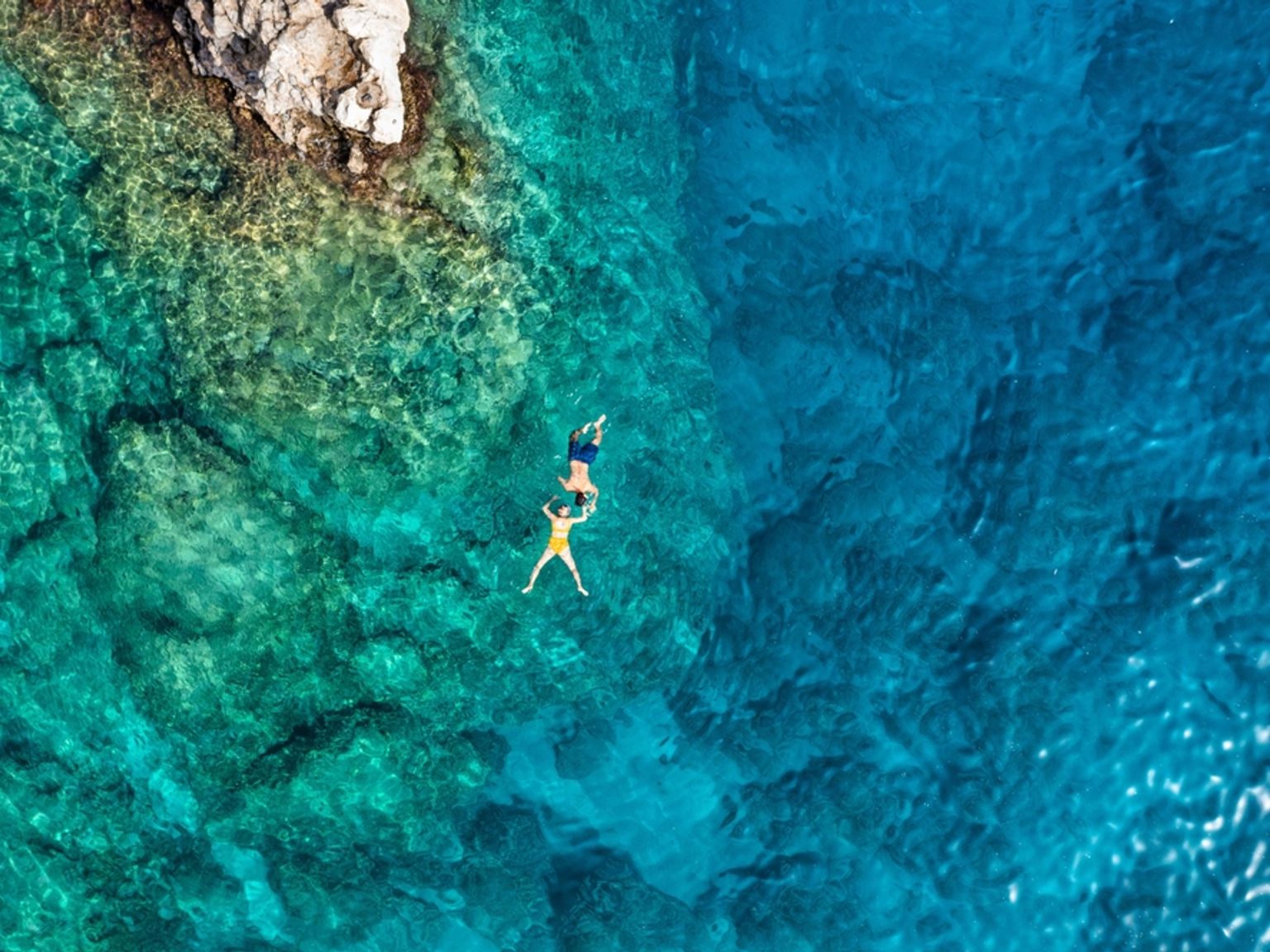 Luftfoto av to personer som flyter i klart blågrønt havvann nær en steinete kystlinje. Rhodos, Hellas