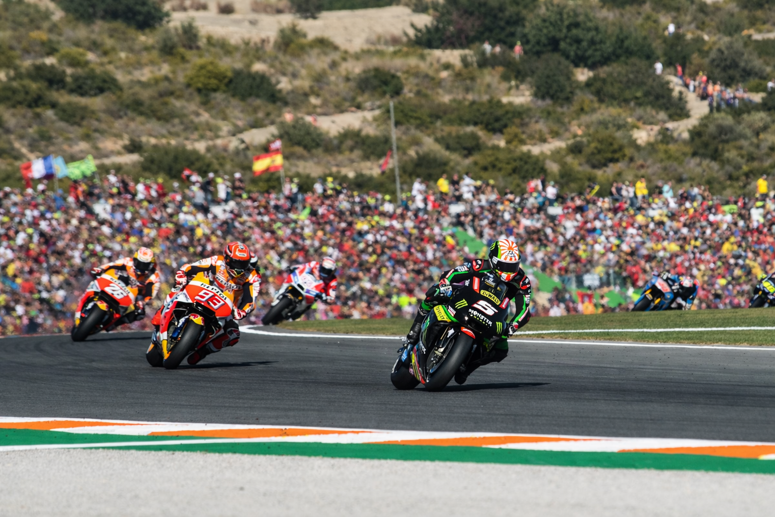 Several motorcycle racers lean into a sharp turn on a track with a large crowd on the hills in the background.