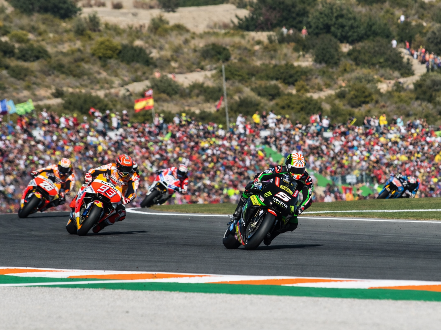 Mehrere Motorradrennfahrer legen sich in eine scharfe Kurve auf einer Rennstrecke, während im Hintergrund eine große Menschenmenge auf den Hügeln zuschaut.