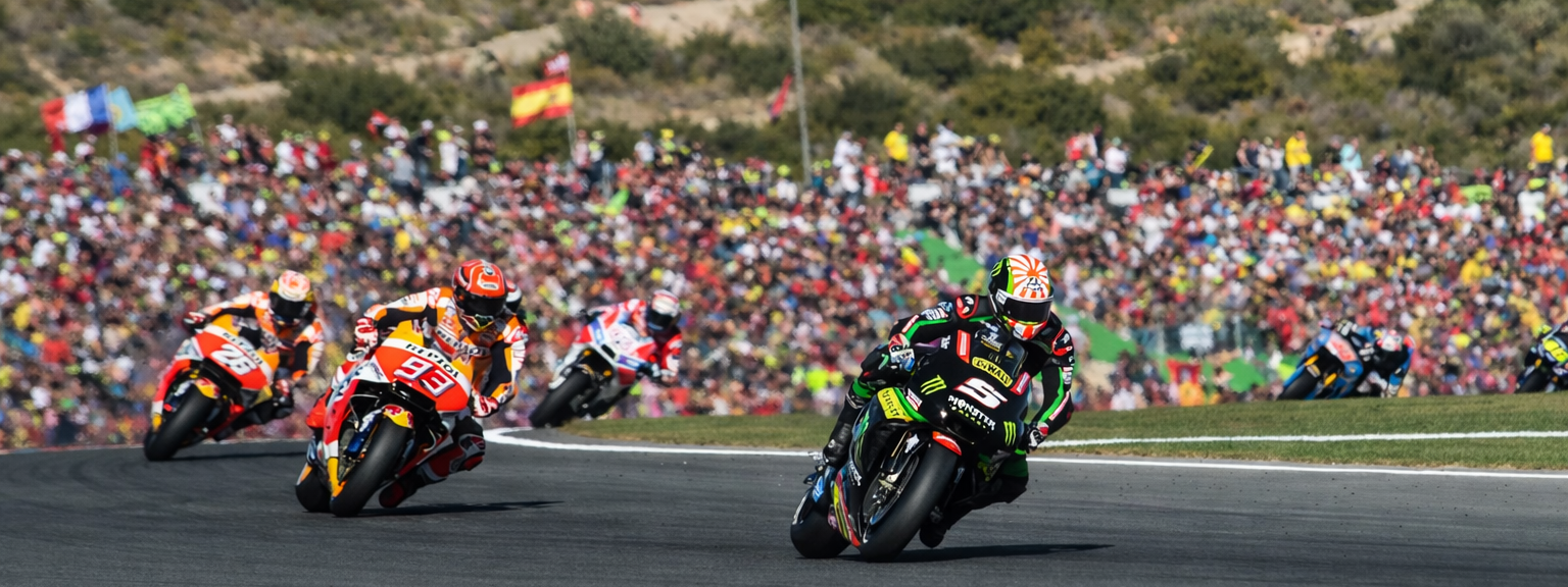 Mehrere Motorradrennfahrer legen sich in eine scharfe Kurve auf einer Rennstrecke, während im Hintergrund eine große Menschenmenge auf den Hügeln zuschaut.