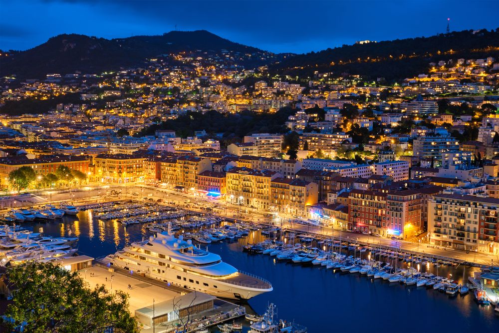 Nighttime aerial view of a vibrant city harbor with numerous boats, illuminated buildings, and city lights stretching up the hills. Nice, France