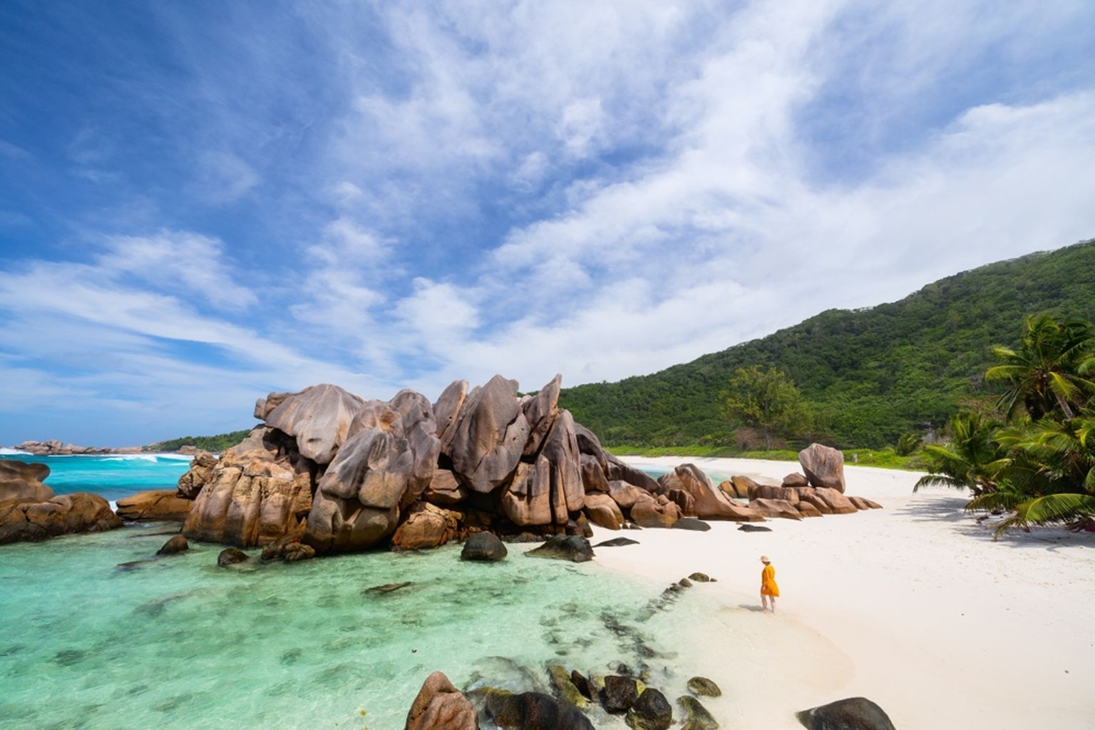 En person i gult går på en hvid sandstrand med store granitblokke og klart turkis vand. Seychellerne