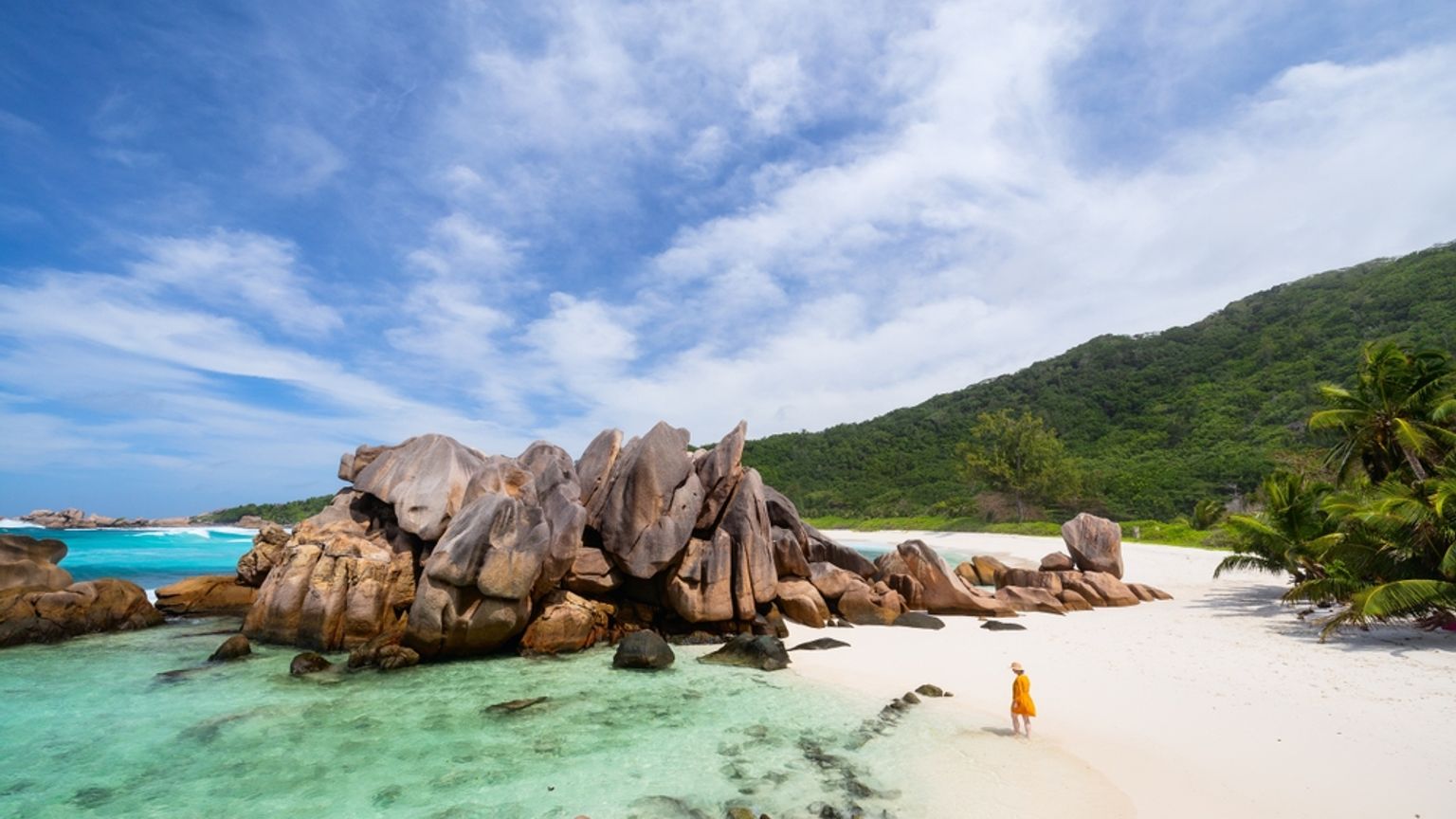 En person i gult går på en hvid sandstrand med store granitblokke og klart turkis vand. Seychellerne