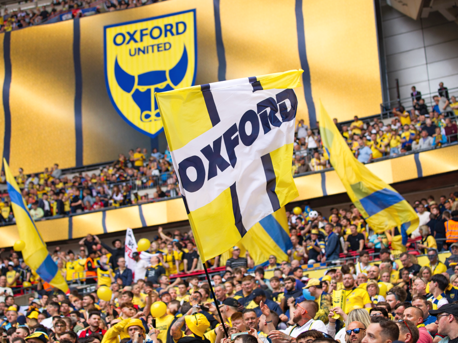 A large crowd of Oxford United fans in a stadium, waving yellow and blue flags, with the team's logo banner in the background.