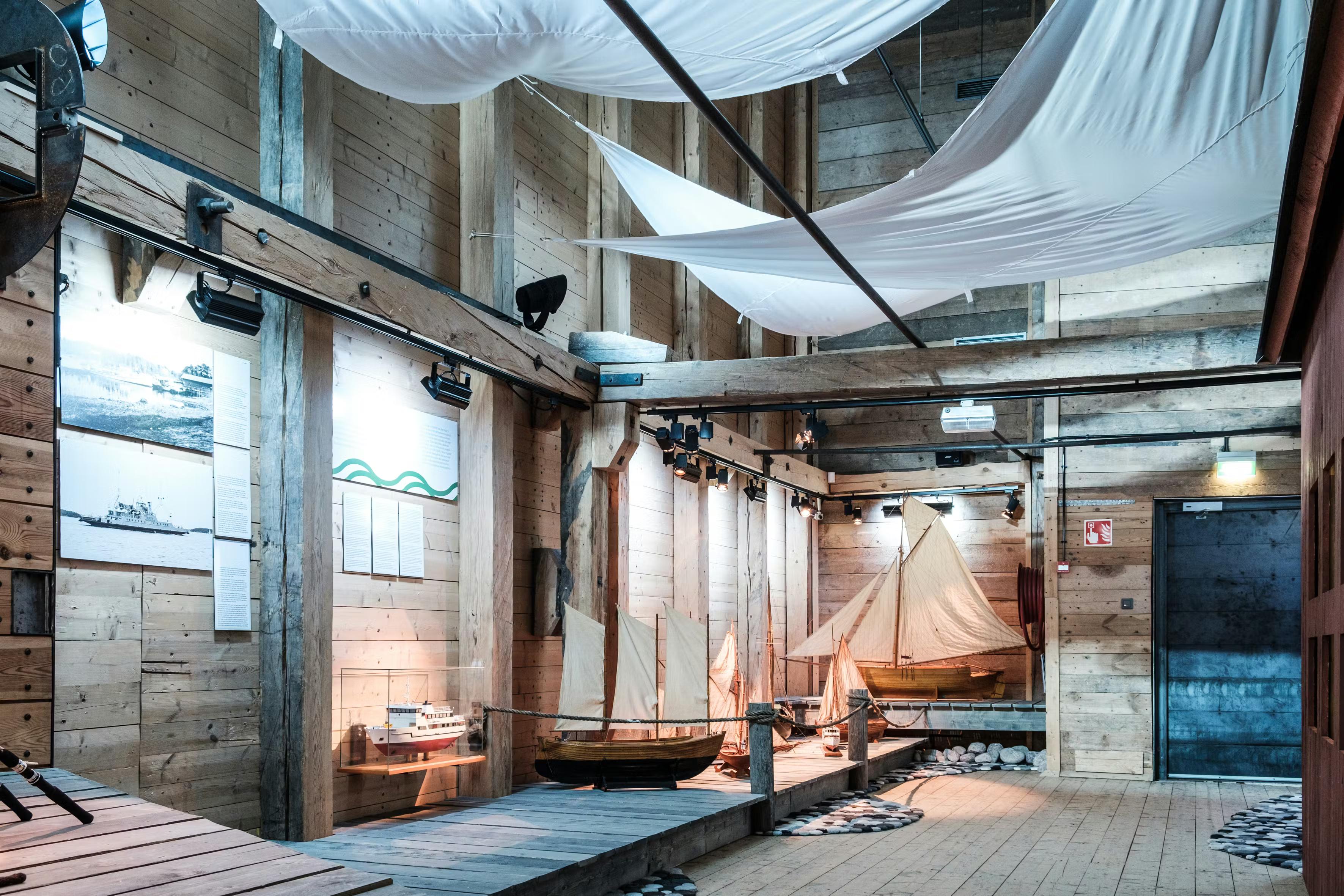 An indoor museum exhibit with model boats on wooden piers, surrounded by wooden walls, and white fabric draped from the ceiling.