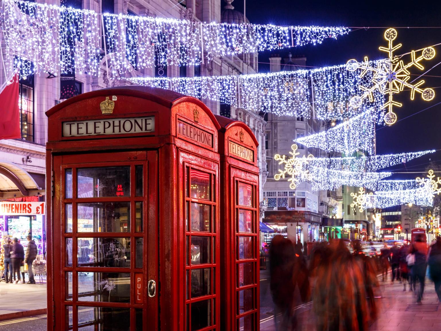 Röda telefonkiosker på en gata i London upplysta med vita ljusslingor och snöflingor på natten, med suddiga fotgängare.