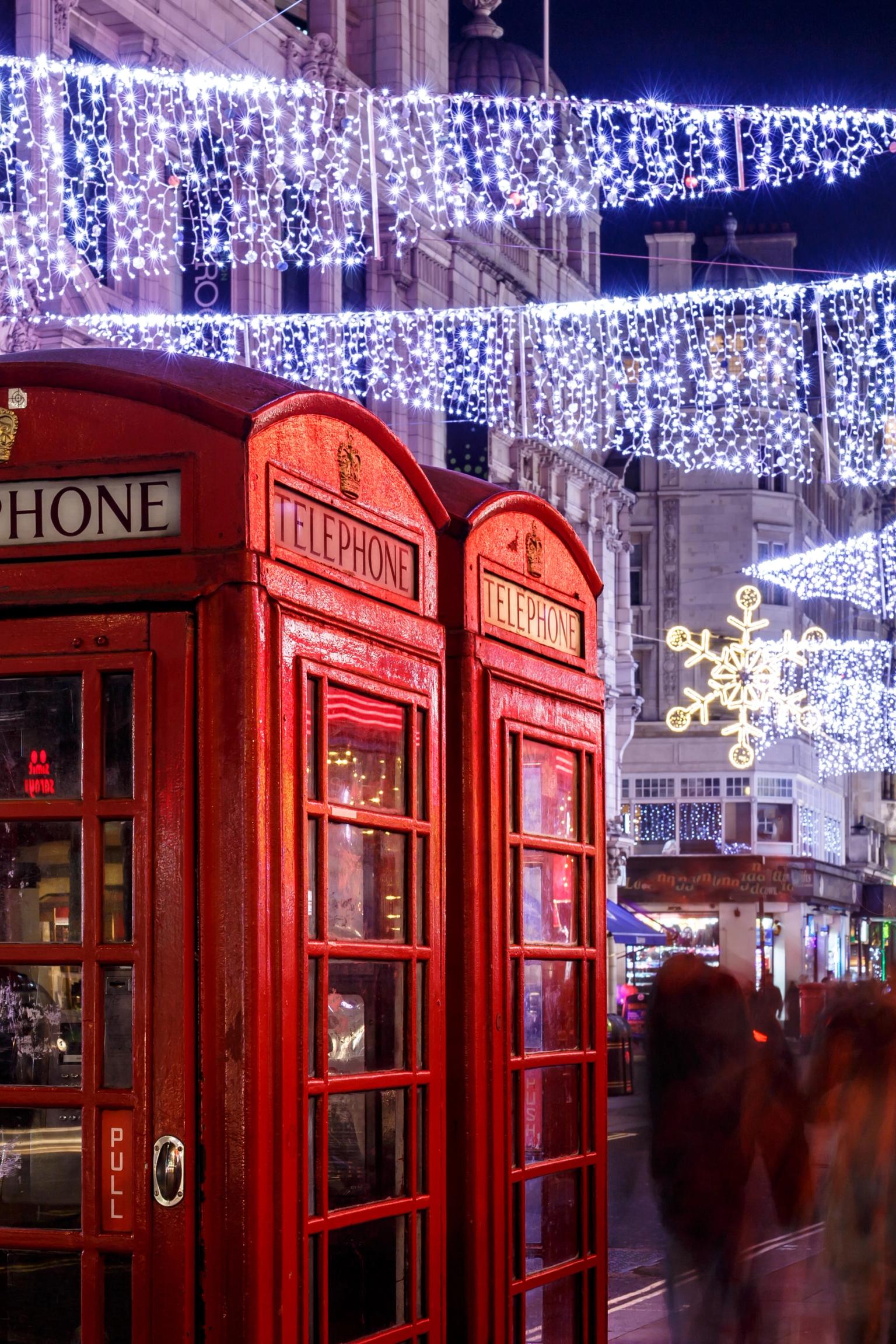 Röda telefonkiosker på en gata i London upplysta med vita ljusslingor och snöflingor på natten, med suddiga fotgängare.
