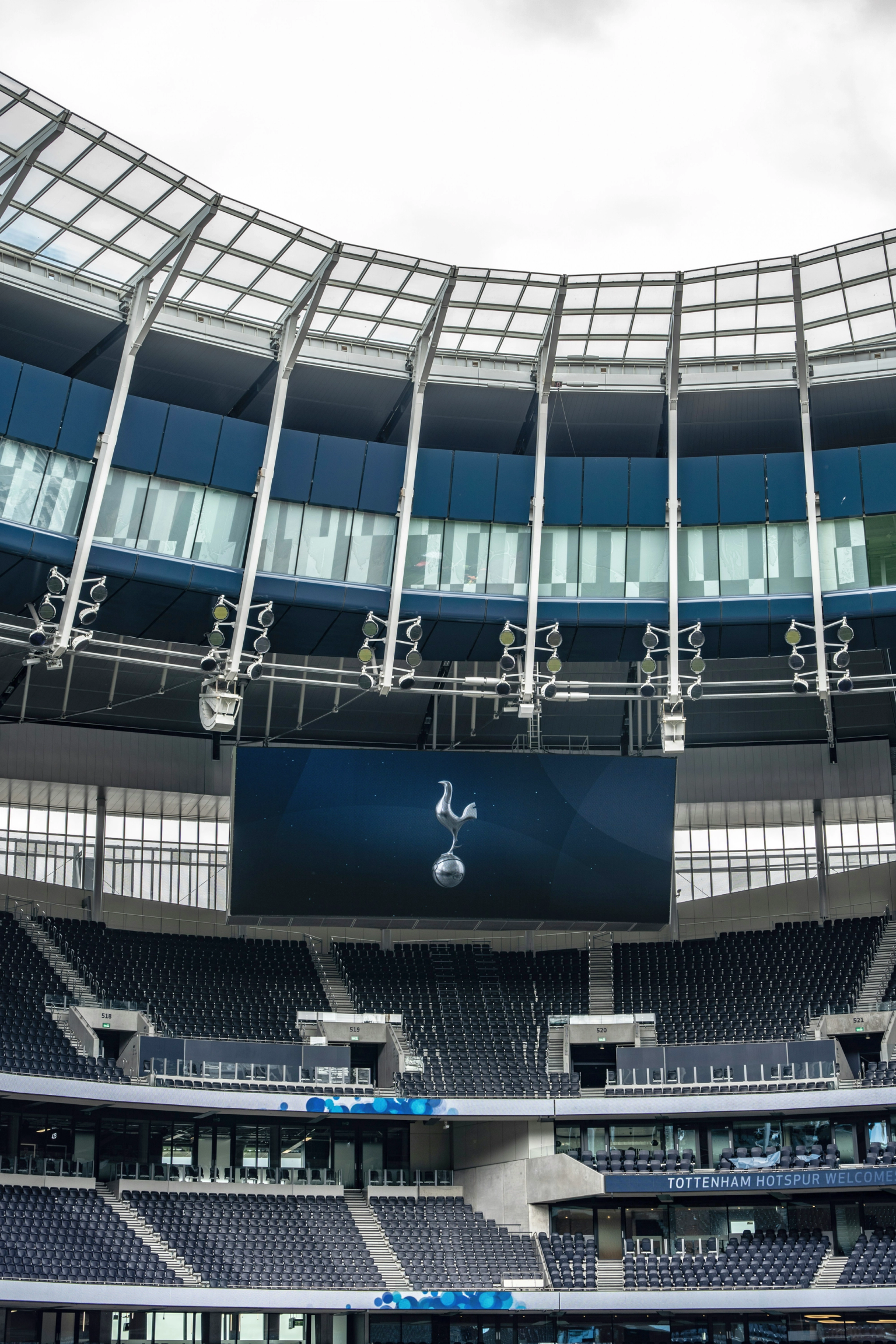 Interiören av Tottenham Hotspur Stadium med mörkblå tomma säten, en stor skärm som visar klubbens tupp-logotyp och en modern takstruktur ovanför.