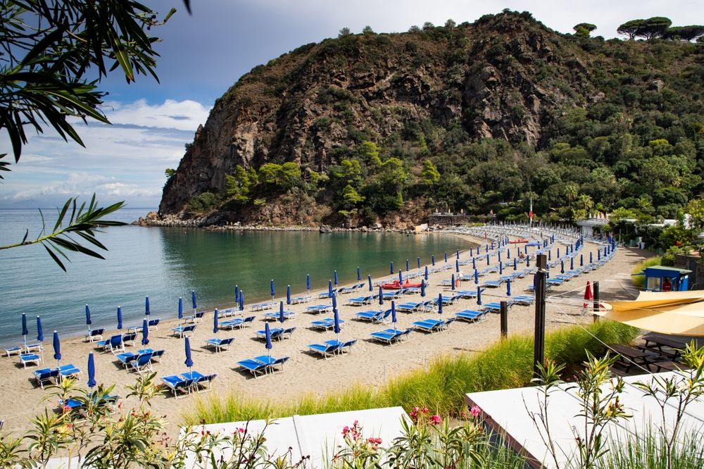 Plage de sable incurvée avec des rangées de chaises longues et de parasols bleus, eau calme et grande falaise boisée. Procida, Italie