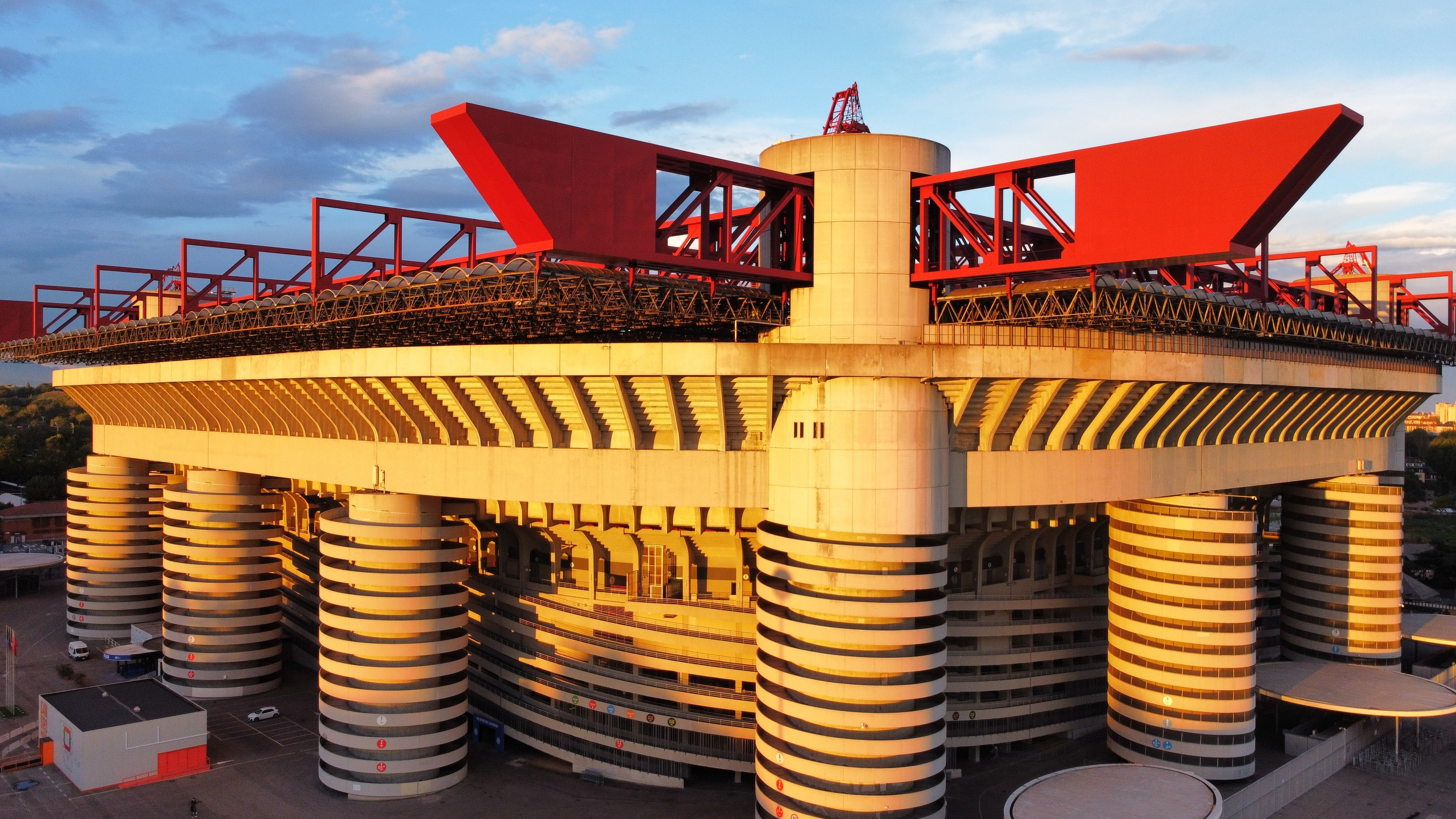 A Brutalist stadium with red roof structures and spiral concrete ramps, illuminated by golden hour sunlight.