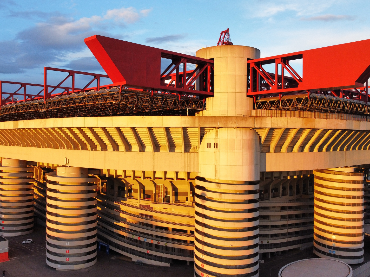 A Brutalist stadium with red roof structures and spiral concrete ramps, illuminated by golden hour sunlight.