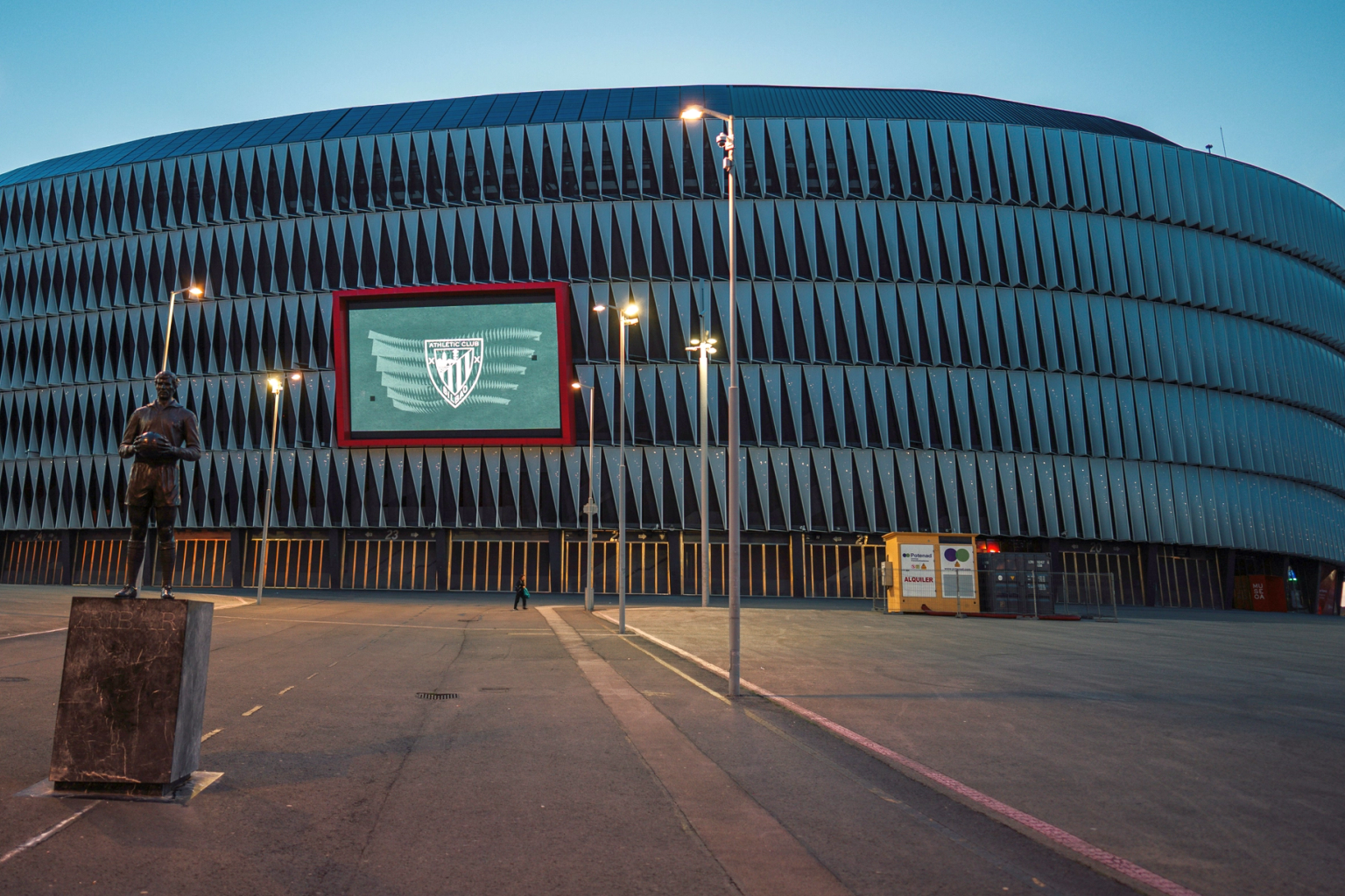 San Mamés Stadion buitenkant bij schemering, met het Athletic Bilbao-logo op een scherm en een spelersstandbeeld.