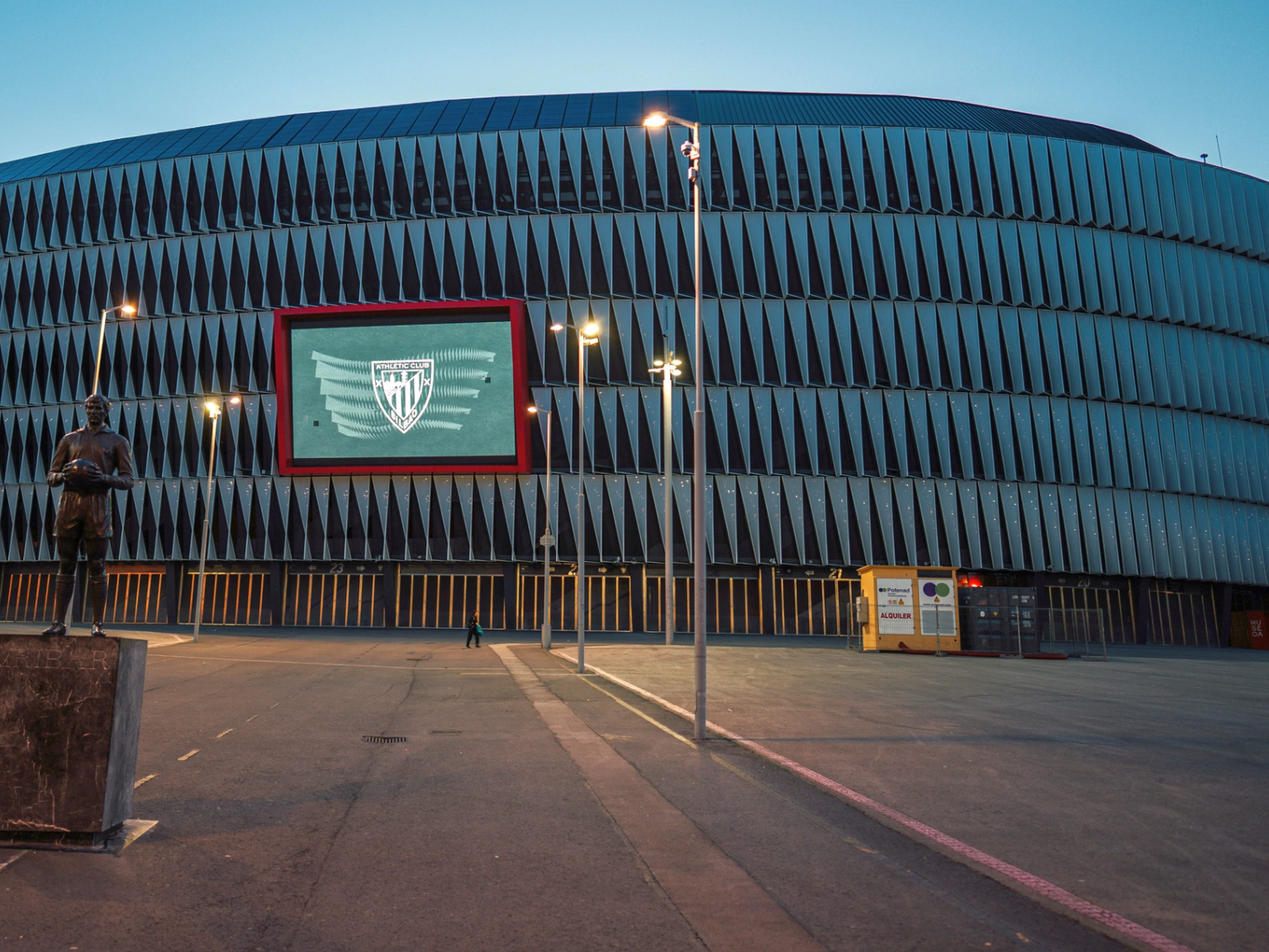 San Mamés-stadionens exteriör i skymningen, med Athletic Bilbaos logotyp på en skärm och en spelarestaty.