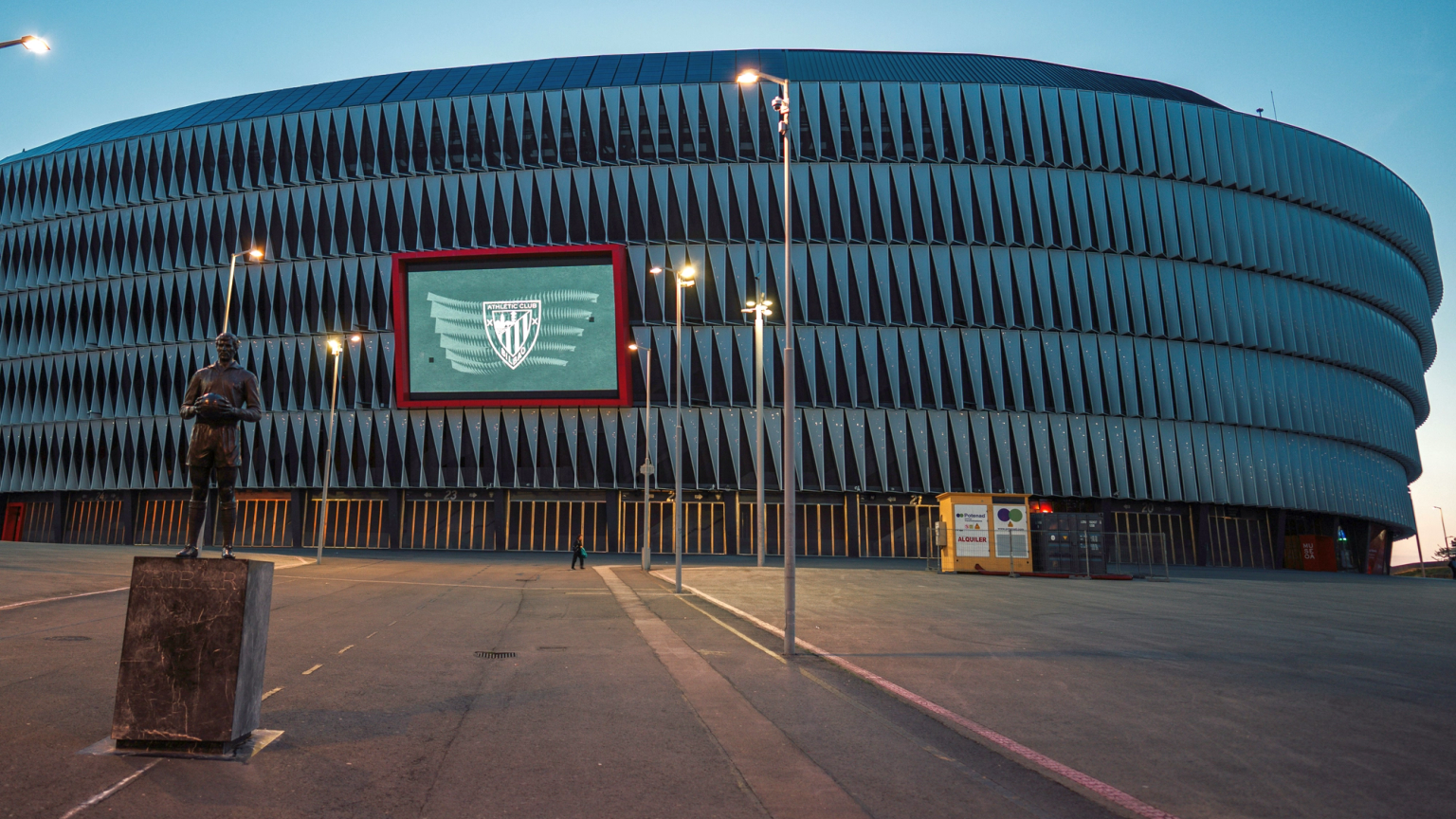 San Mamés Stadion buitenkant bij schemering, met het Athletic Bilbao-logo op een scherm en een spelersstandbeeld.