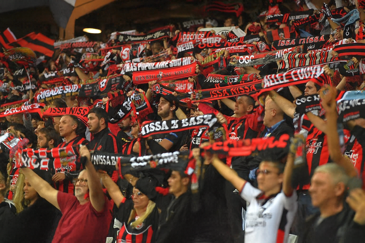 Uma multidão de torcedores de futebol aplaudindo, segurando cachecóis pretos e vermelhos em um estádio.