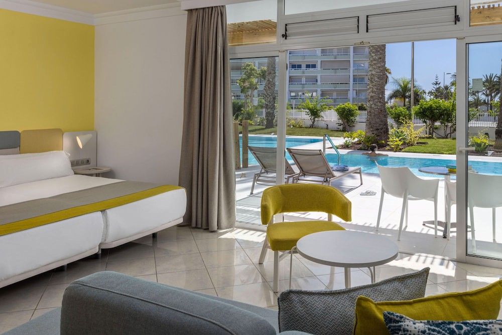Et moderne hotelværelse med en seng og siddeområde, med en gul accentvæg og direkte adgang til en udendørs swimmingpool og have.