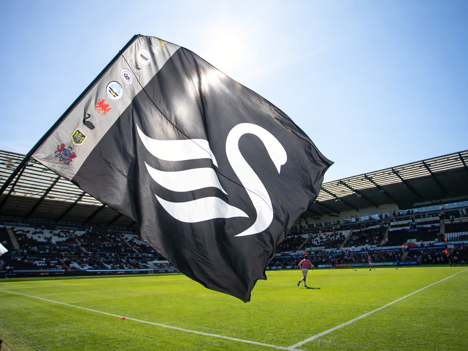 Black Swansea City AFC flag with a white swan logo flying in a sunny football stadium.