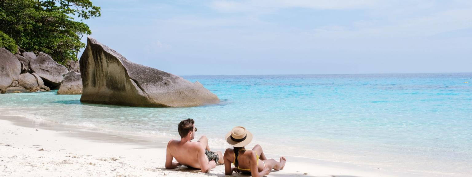 Ett par går hand i hand på en vit sandstrand med turkost vatten och en lutande palm.