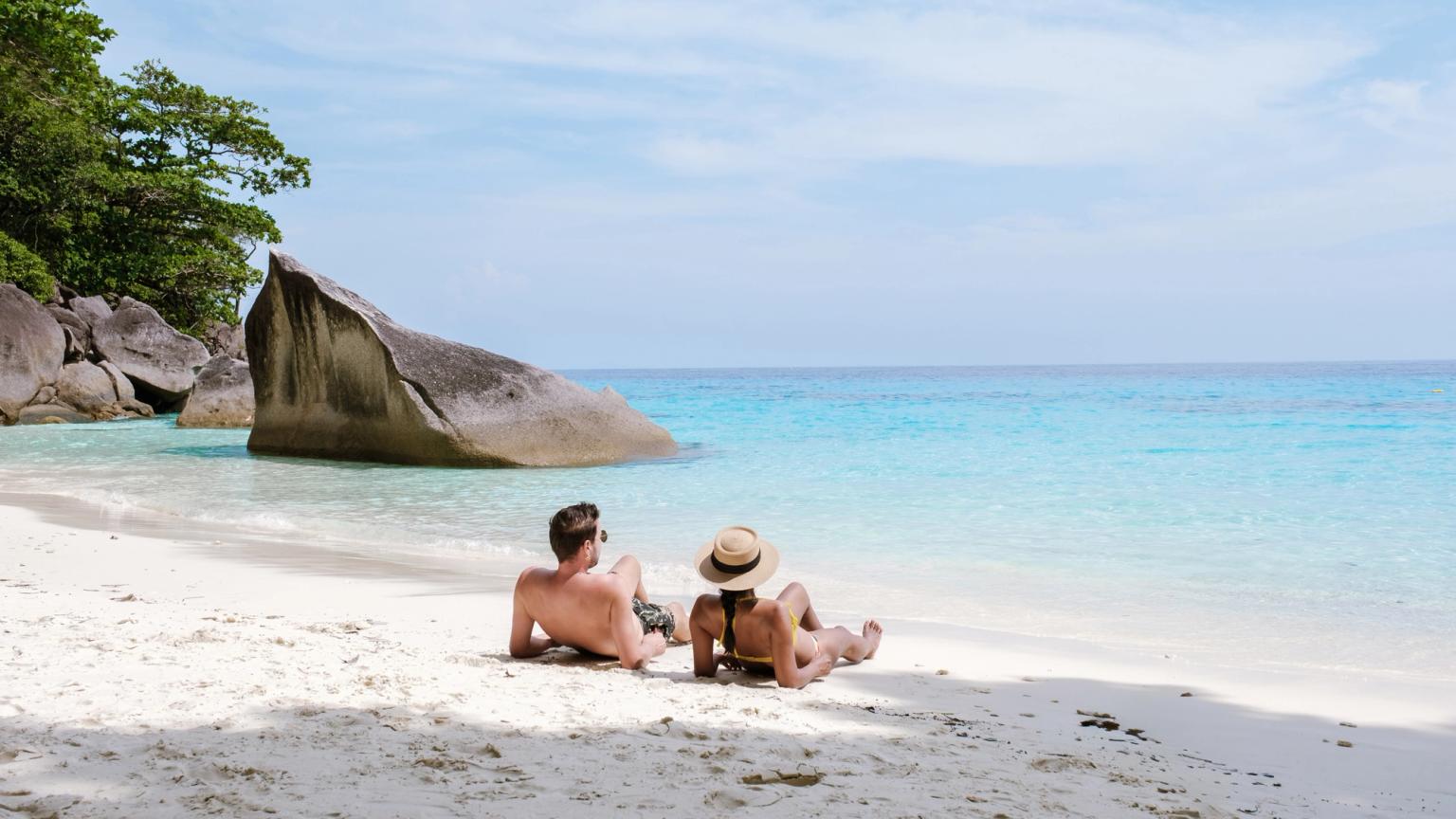 Ett par går hand i hand på en vit sandstrand med turkost vatten och en lutande palm.