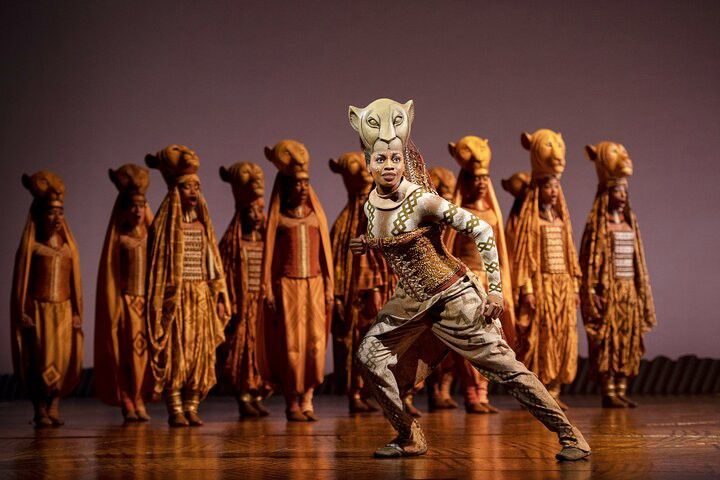 A performer from The Lion King musical in an elaborate lioness costume and mask strikes an active pose on stage, with other performers in golden costumes behind.