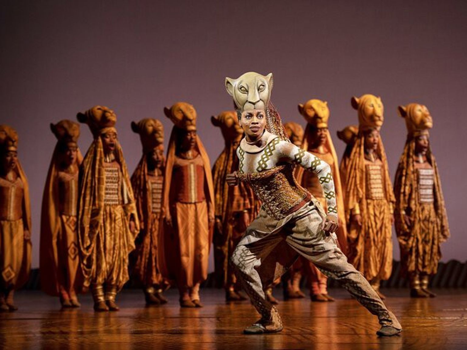 A performer from The Lion King musical in an elaborate lioness costume and mask strikes an active pose on stage, with other performers in golden costumes behind.