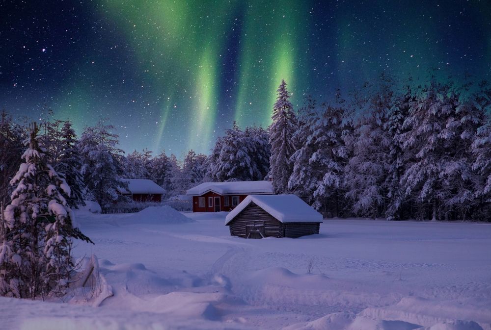 En snöig vinterby på natten, med röda och trästugor inbäddade bland snötäckta träd, under en stjärnklar himmel upplyst av livliga gröna norrsken. Rovaniemi