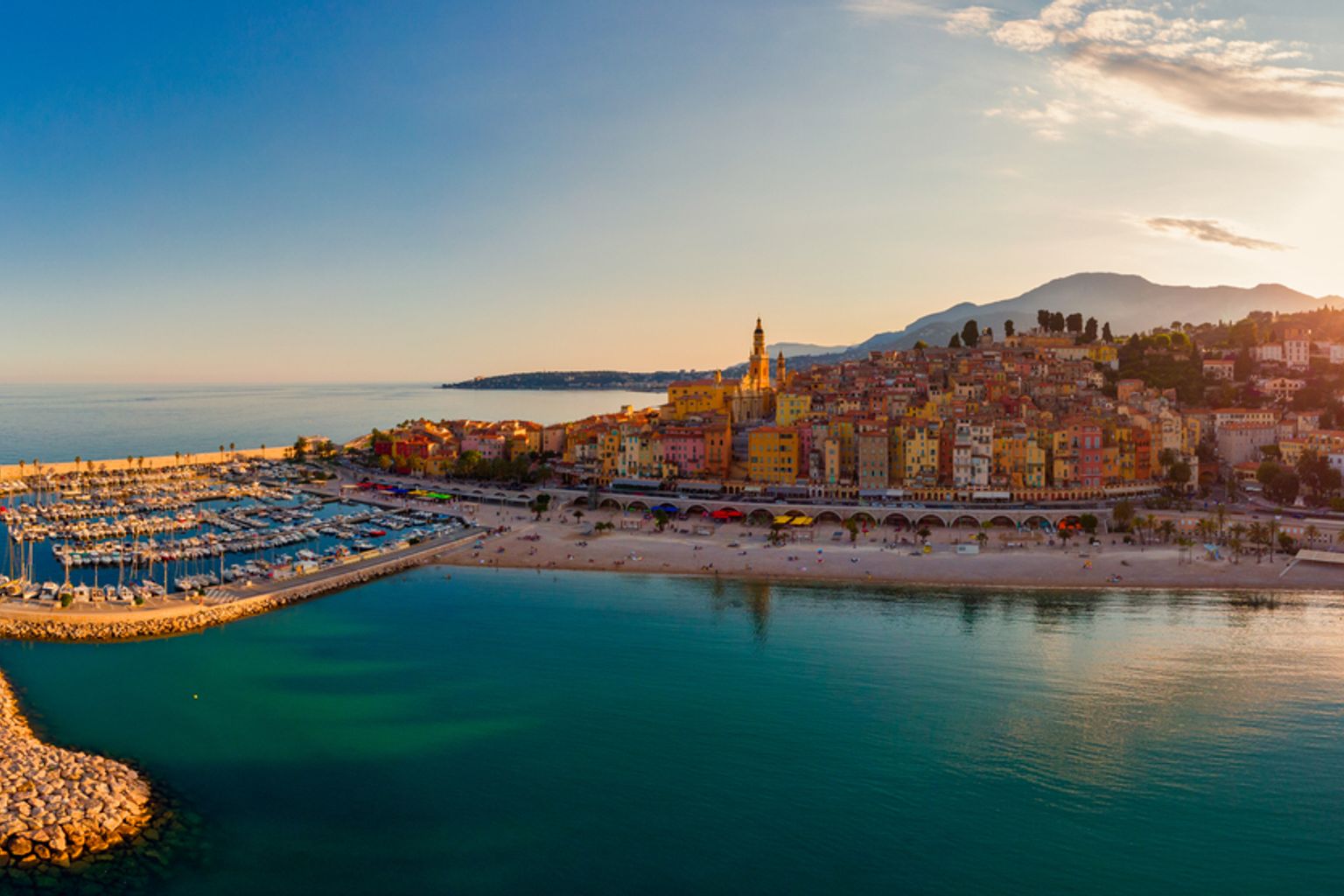 Flygvy över en livlig kuststad med en marina, strand och färgglada byggnader på sluttningarna under varmt kvällsljus. Menton, Frankrike.