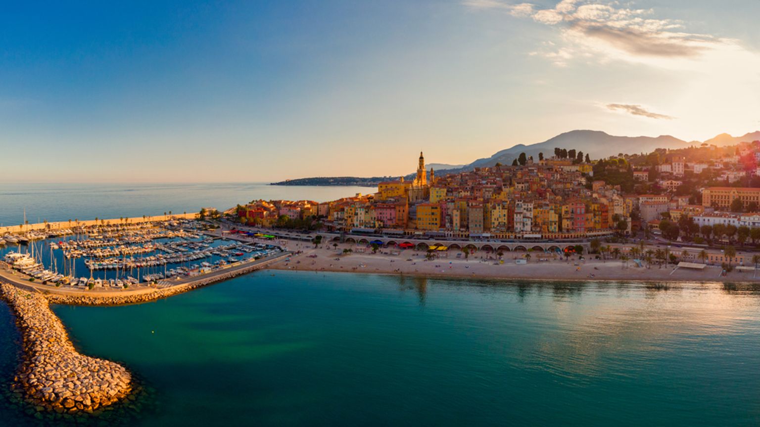 Flygvy över en livlig kuststad med en marina, strand och färgglada byggnader på sluttningarna under varmt kvällsljus. Menton, Frankrike.