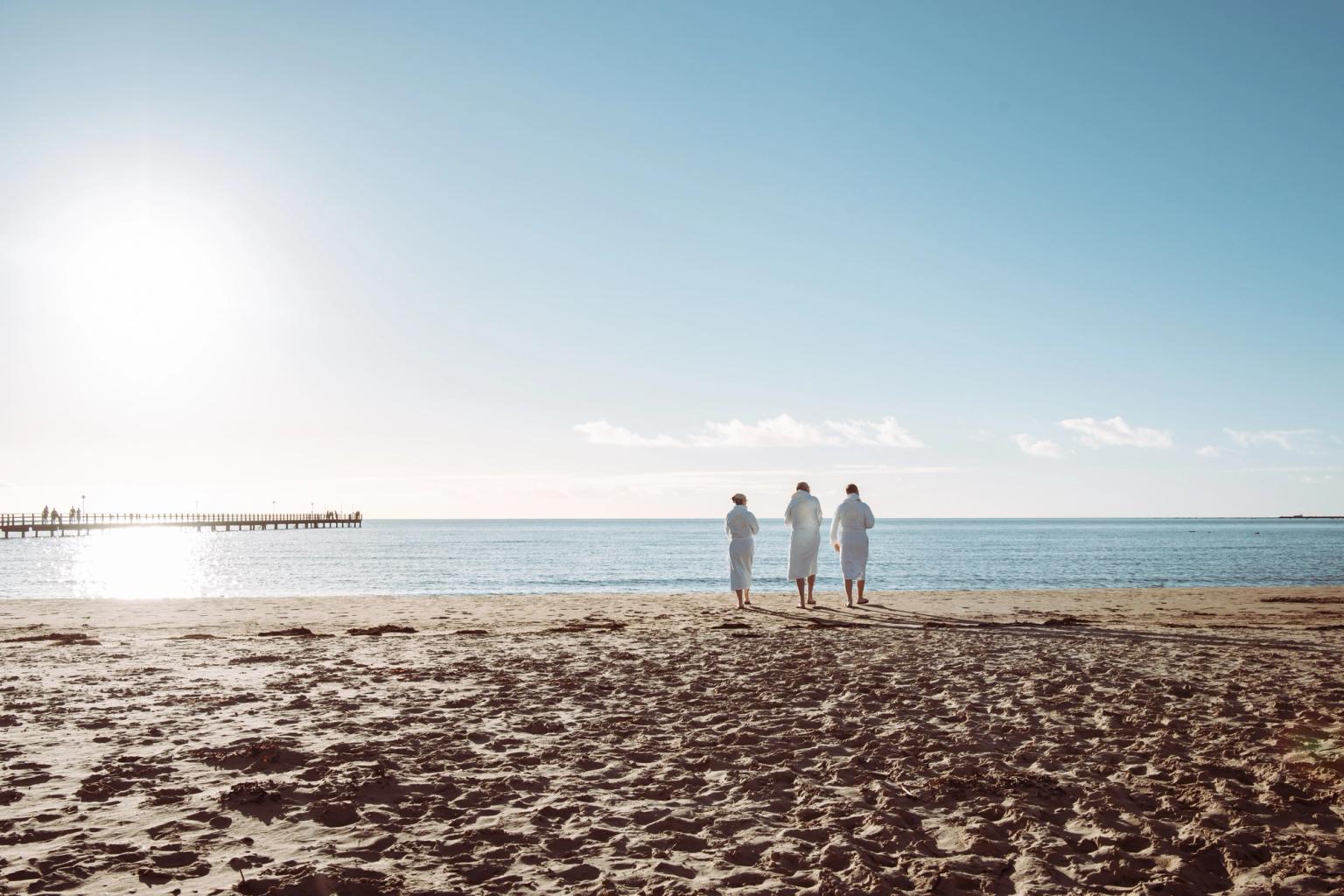Tre personer i vita badrockar står på en solig strand, vända mot havet med en pir i fjärran.