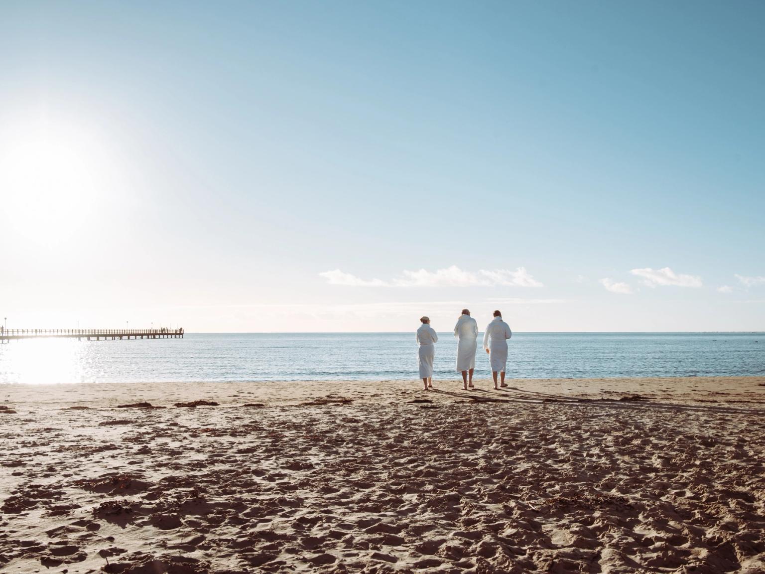 Tre personer i hvide badekåber står på en solrig strand med ansigtet vendt mod havet og en mole i det fjerne.