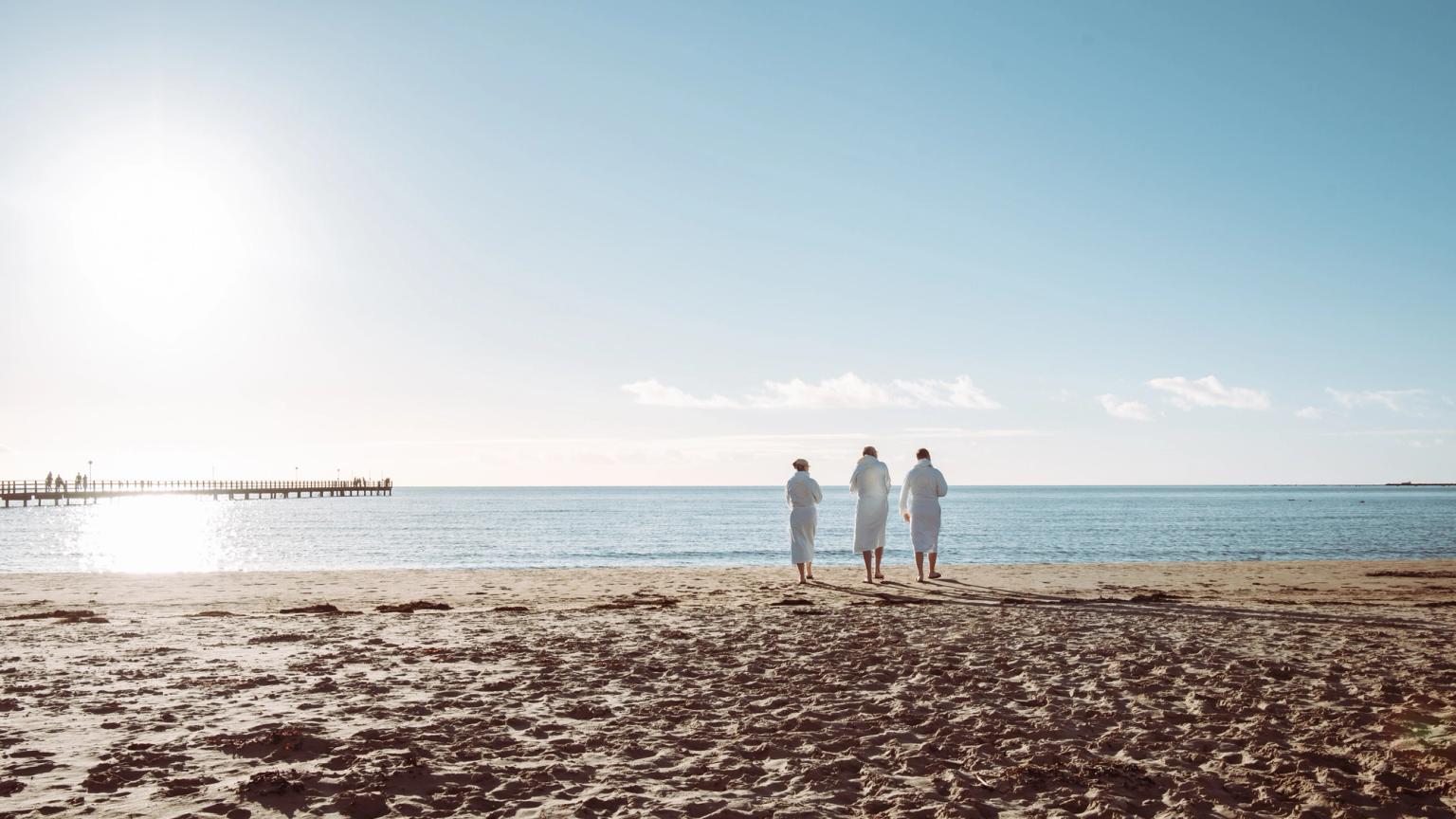 Tre personer i vita badrockar står på en solig strand, vända mot havet med en pir i fjärran.