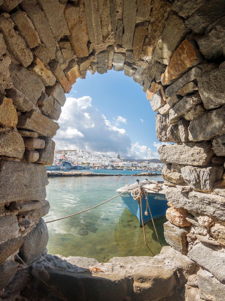 A blue boat in clear water viewed through a stone archway, with a white village and cloudy sky in the background. Paros, Greece