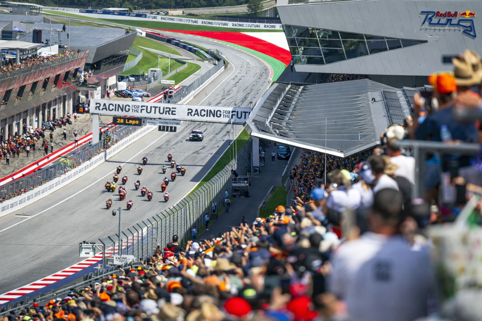 Motorbiciklik versenyeznek a Red Bull Ring versenypályáján, miközben a nézők megtöltik a lelátókat.