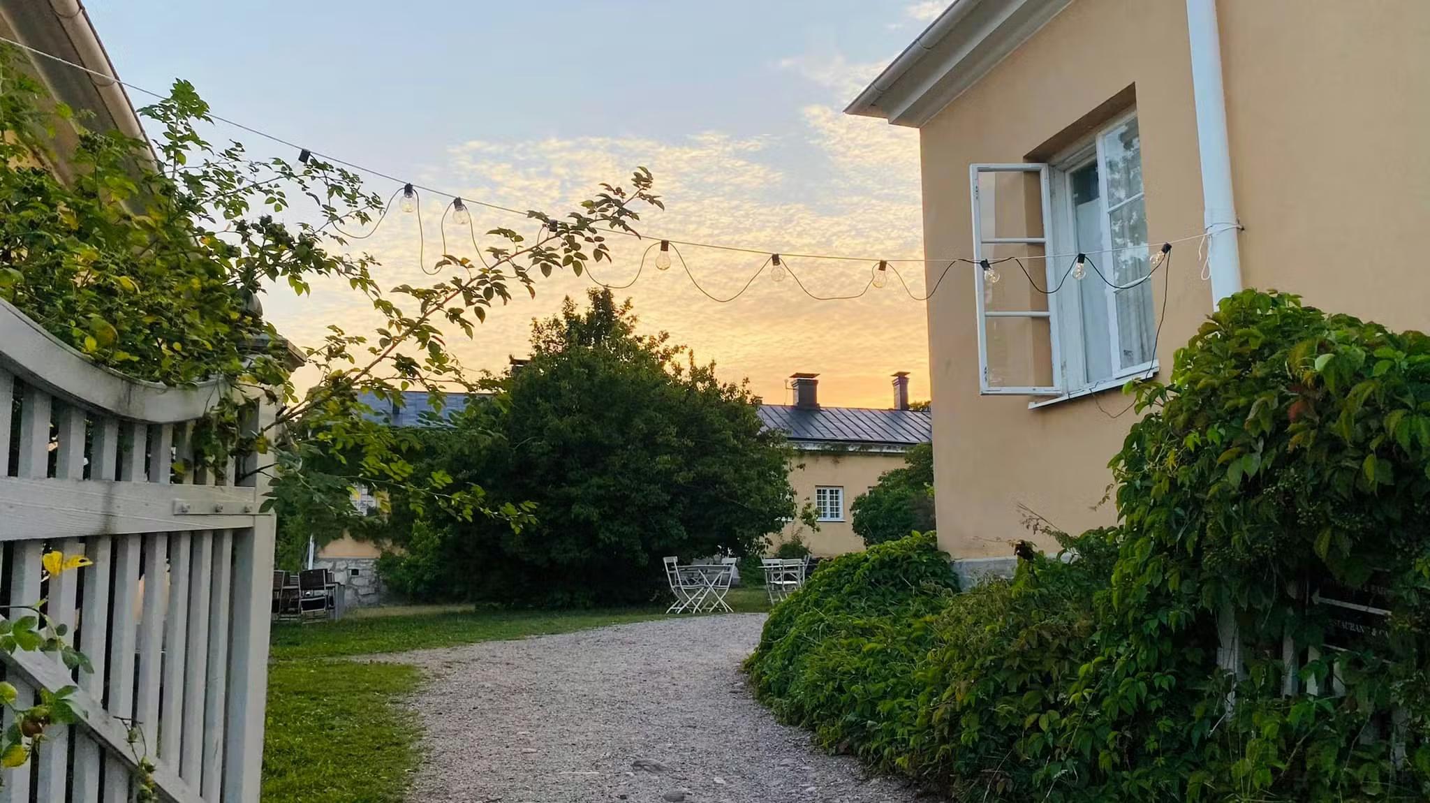 Charming garden path with string lights, a yellow building, and green foliage at sunset.