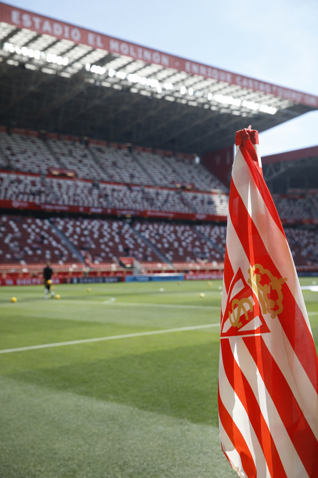 A red and white striped corner flag with a gold crest stands in the foreground of a football stadium with empty red and white seats and a green field.