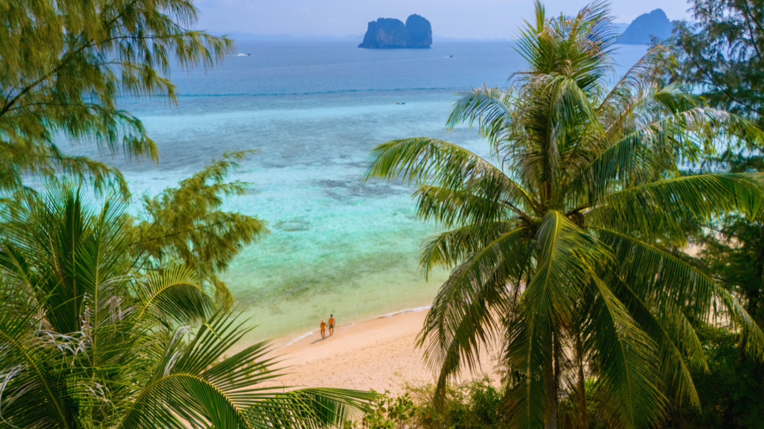 View of a tropical beach with turquoise water, two people walking, distant islands, and framing palm trees.