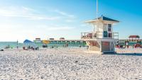 Hengenpelastajan torni valkoisella hiekkarannalla, jossa on laituri, ihmisiä ja sininen taivas. Clearwater Beach, Florida