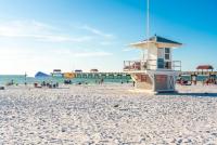 Hengenpelastajan torni valkoisella hiekkarannalla, jossa on laituri, ihmisiä ja sininen taivas. Clearwater Beach, Florida