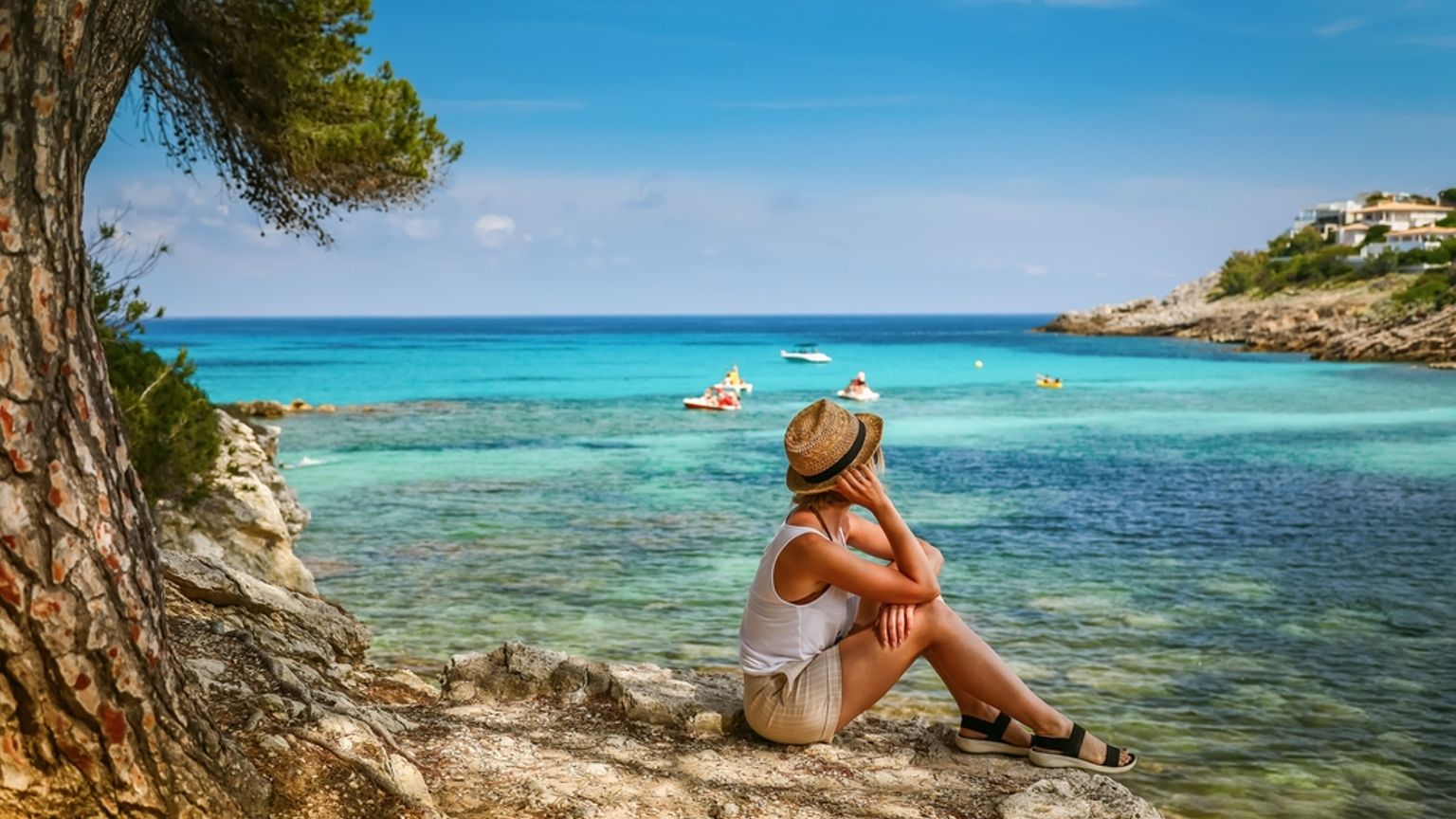 En kvinna med stråhatt sitter på klippor och blickar ut över en ljus turkos vik med båtar. Mallorca, Spanien.