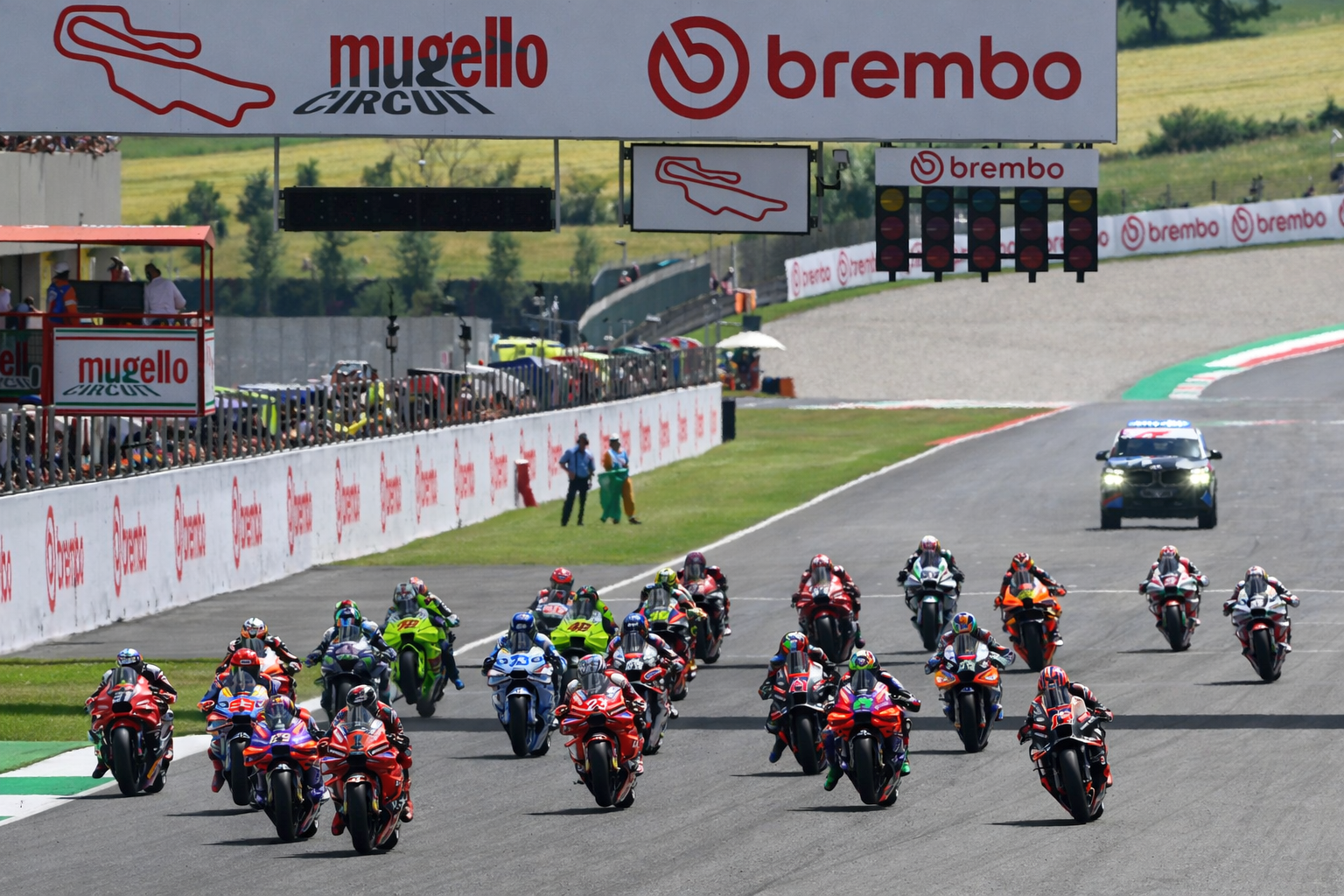 Motorsykler racer nedover Mugello Circuit-banen med Brembo-bannere og startlys synlige.
