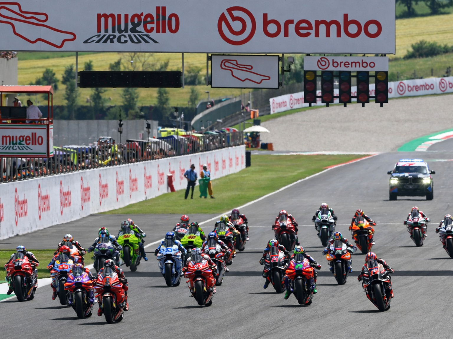 Motorsykler racer nedover Mugello Circuit-banen med Brembo-bannere og startlys synlige.
