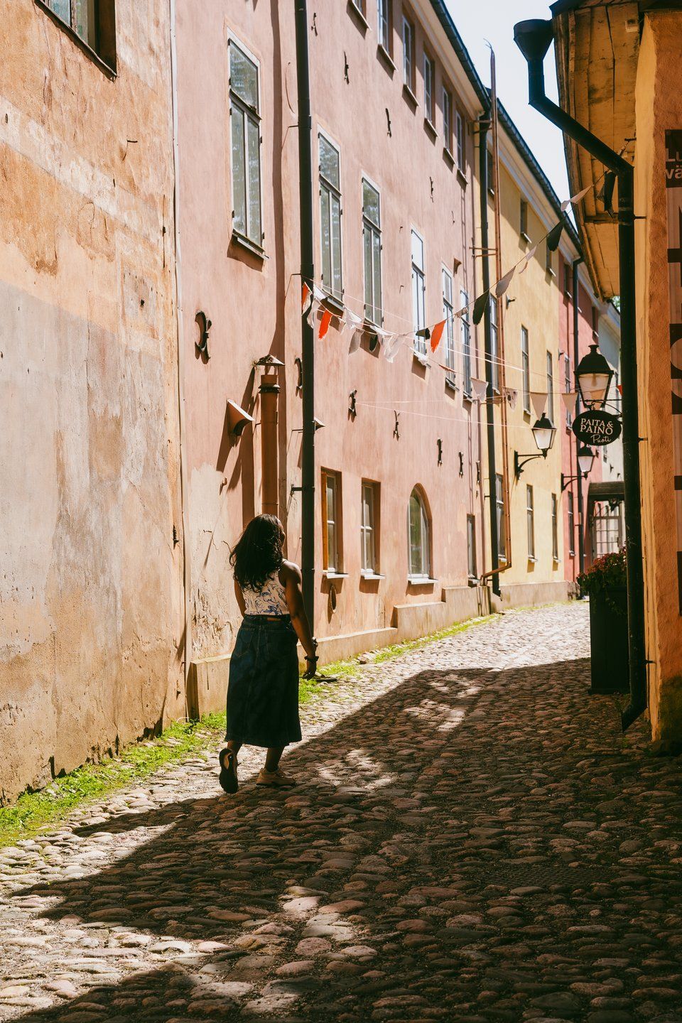 En kvinde går væk på en solrig brostensgade omgivet af farverige bygninger og flag.